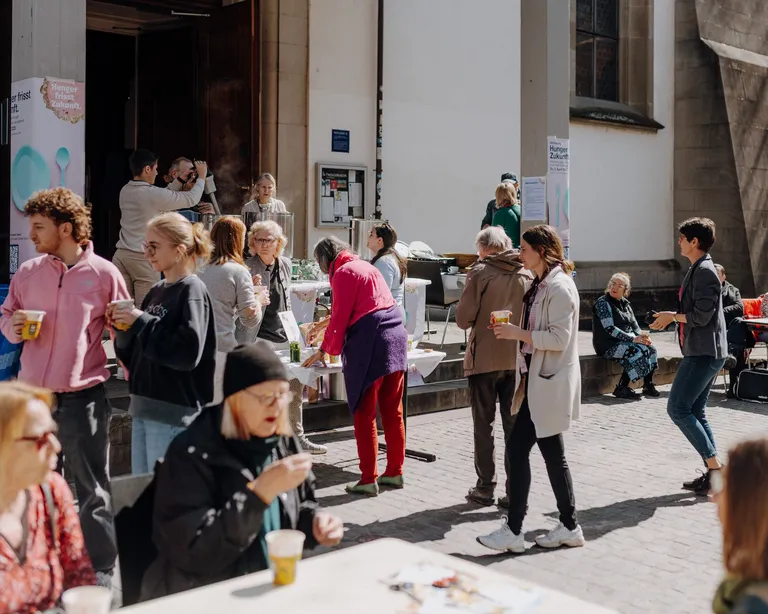 Mehrere Menschen sitzen und stehen bei sonnigem Wetter an Tischen im Freien vor einem Gebäude, unterhalten sich und essen.