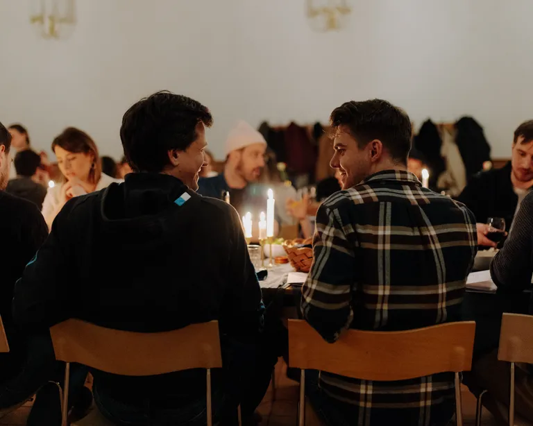 Mehrere Menschen sitzen gemeinsam an einem Tisch in einem festlich beleuchteten Raum und unterhalten sich.