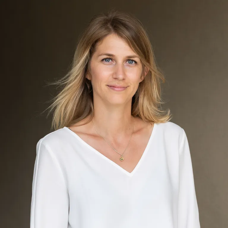 Eine Frau mit schulterlangem, blondem Haar trägt eine weiße Bluse und blickt neutral in die Kamera.