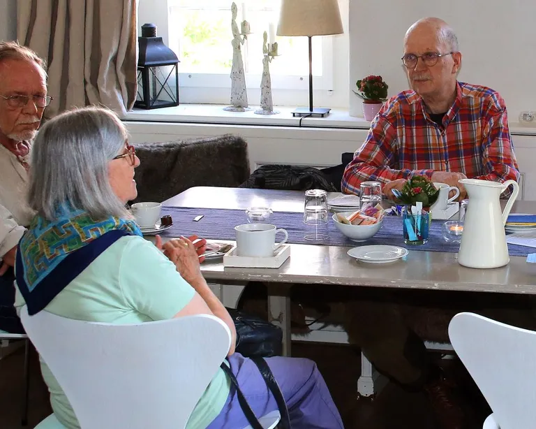 Drei Menschen sitzen an einem Tisch im Wohnzimmer und unterhalten sich bei Kaffee und Gebäck.