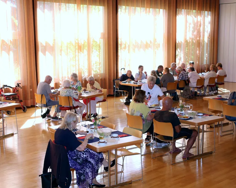 Mehrere ältere Menschen sitzen in einem hellen Raum an Tischen und unterhalten sich oder spielen gemeinsam Spiele.