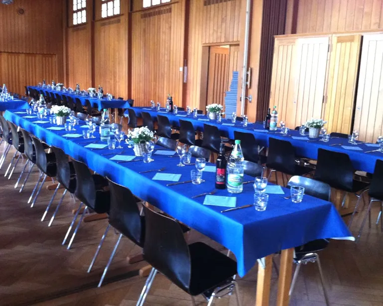 Lange Tische mit blauen Tischdecken, Gläsern und Flaschen in einem holzgetäfelten Saal, bereit für ein Fest.