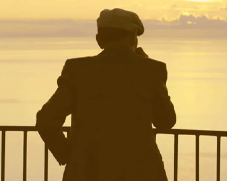 Eine Person mit Hut steht an einem Geländer und blickt auf das Meer bei Sonnenuntergang.