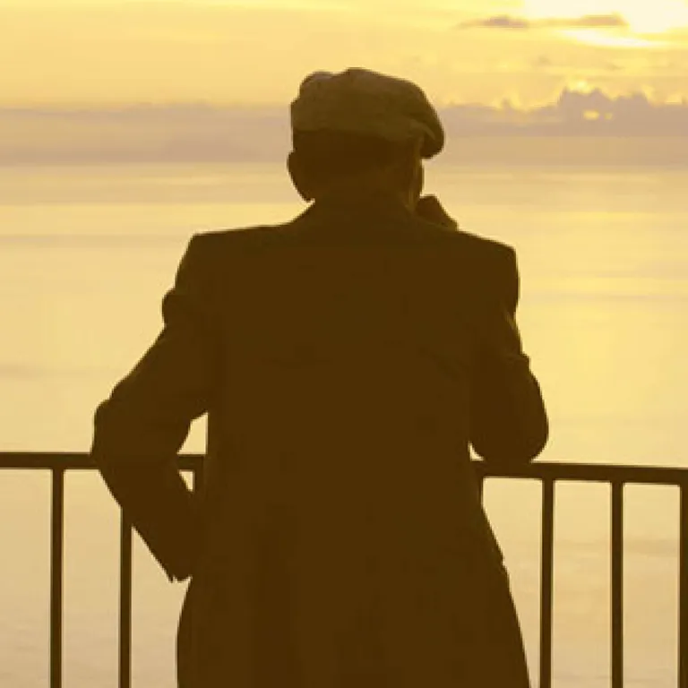 Eine Person mit Hut steht an einem Geländer und blickt auf das Meer bei Sonnenuntergang.