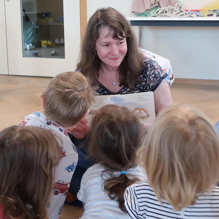 Rebekka Gantenbein liest vier kleinen Kindern in einem Klassenraum ein Buch vor. Alle sitzen auf dem Boden und hören gespannt zu.