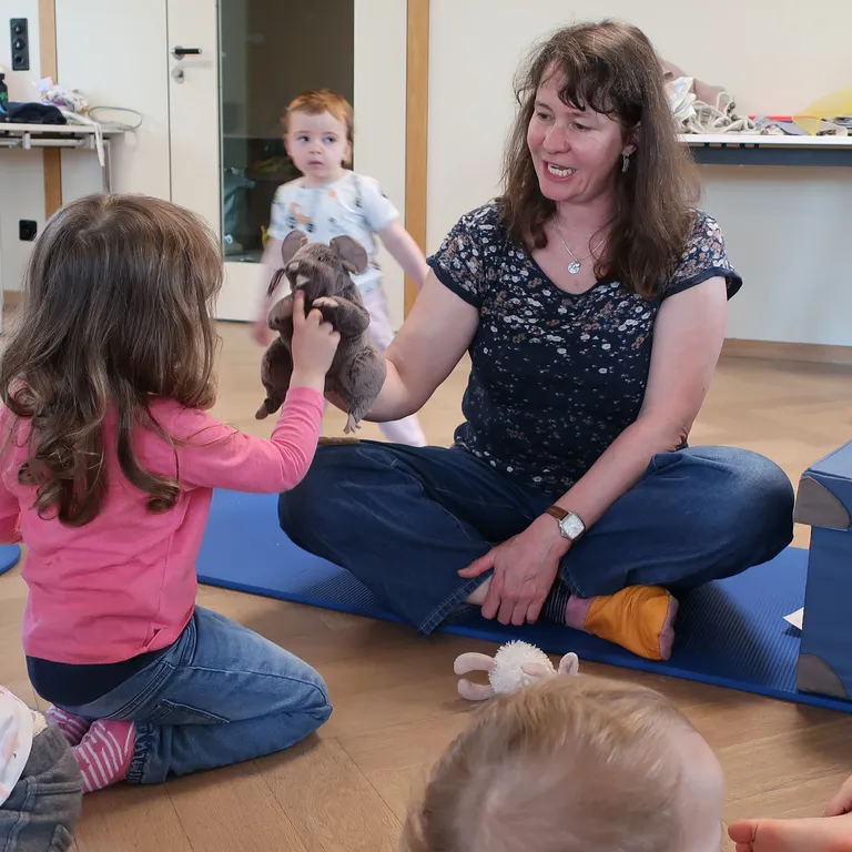Rebekka Gantenbein sitzt mit Kindern auf dem Boden. Sie hält ein Kuscheltier, das sie einem Mädchen im pinken Pullover zeigt.