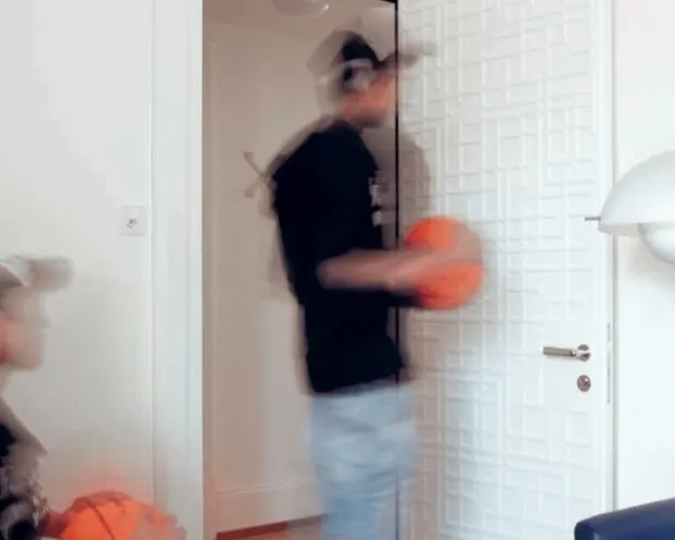 Zwei unscharfe Personen mit einem Basketball in einem Zimmer, eine steht, die andere sitzt oder kauert links im Bild.