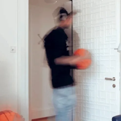 Zwei unscharfe Personen mit einem Basketball in einem Zimmer, eine steht, die andere sitzt oder kauert links im Bild.