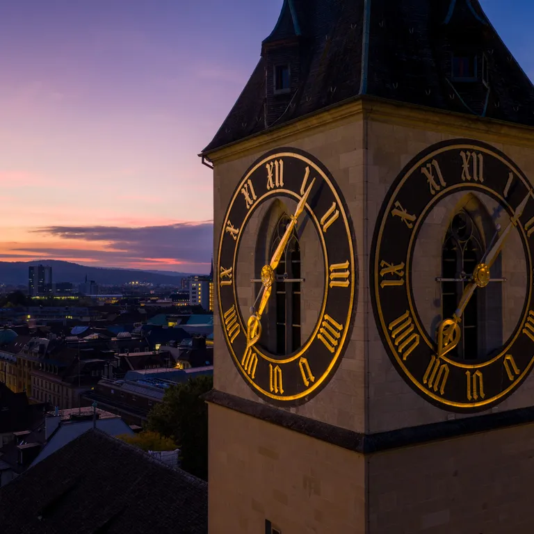 Turmuhr mit goldenen Zifferblättern im Abendlicht, Stadt und Sonnenuntergang im Hintergrund.