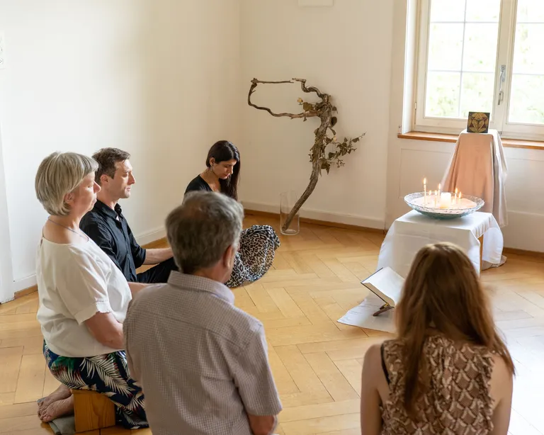 Fünf Personen sitzen meditierend auf Kissen in einem hellen Raum, vor einem kleinen Altar mit Tüchern und Ästen.