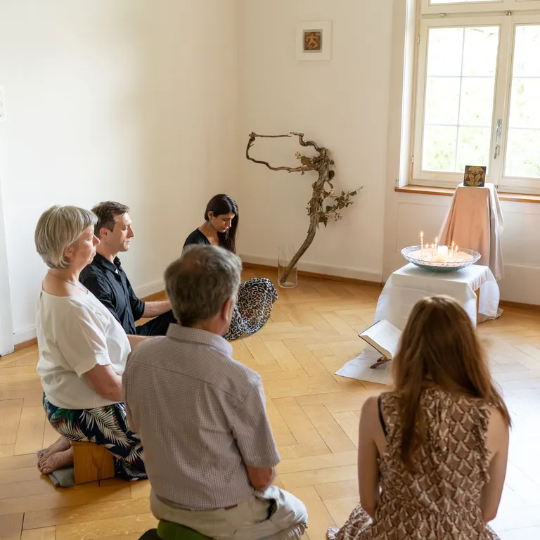 Fünf Personen sitzen meditierend auf Kissen in einem hellen Raum, vor einem kleinen Altar mit Tüchern und Ästen.