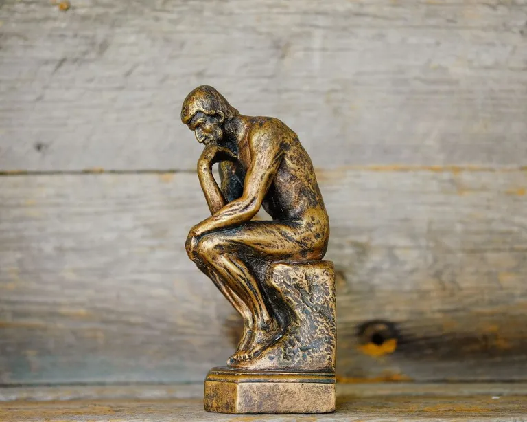 Bronzefarbene Statue eines nachdenklichen Mannes, der den Kopf auf die Hand stützt, vor Holzhintergrund.