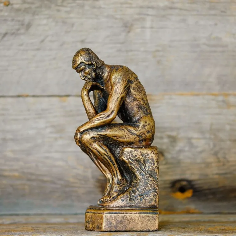 Bronzefarbene Statue eines nachdenklichen Mannes, der den Kopf auf die Hand stützt, vor Holzhintergrund.
