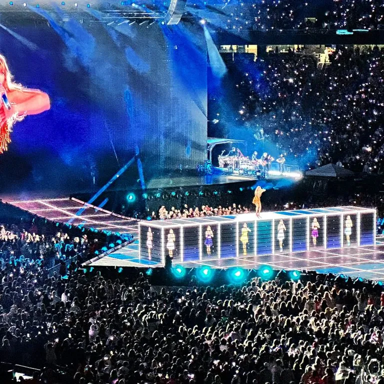 Eine Sängerin tritt auf einer grossen, hell erleuchteten Bühne in einem vollen Stadion auf, mit Leinwand im Hintergrund.