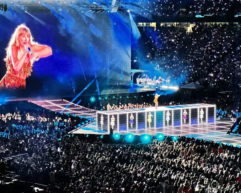 Eine Sängerin tritt auf einer grossen, hell erleuchteten Bühne in einem vollen Stadion auf, mit Leinwand im Hintergrund.