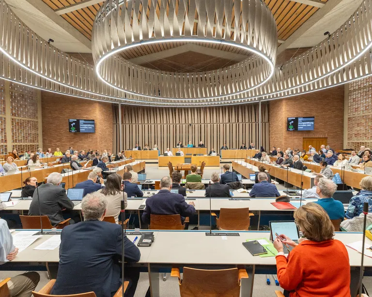 Großer Konferenzsaal, viele Menschen sitzen an Tischen und hören einem Vortrag oder einer Diskussion zu.