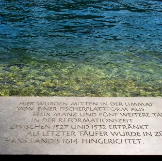 Helle Gedenktafel mit Inschrift am Ufer eines Flusses, im Hintergrund klares, grünbläuliches Wasser und Kieselsteine sichtbar.