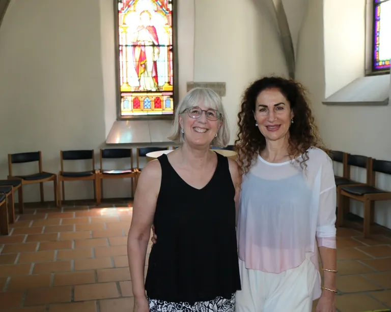 Zwei Frauen stehen in einem hellen Raum mit Stühlen und buntem Kirchenfenster im Hintergrund.