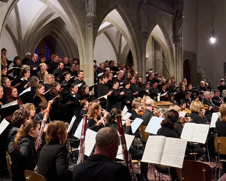 Großes Orchester und Chor musizieren gemeinsam in einer Kirche mit gotischen Bögen im Hintergrund.