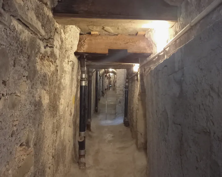 Schmaler, alter Tunnel mit Beton- und Steinwänden, schwach beleuchtet durch mehrere Lampen an der Decke.