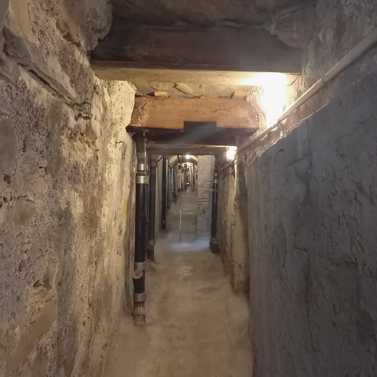 Schmaler, alter Tunnel mit Beton- und Steinwänden, schwach beleuchtet durch mehrere Lampen an der Decke.