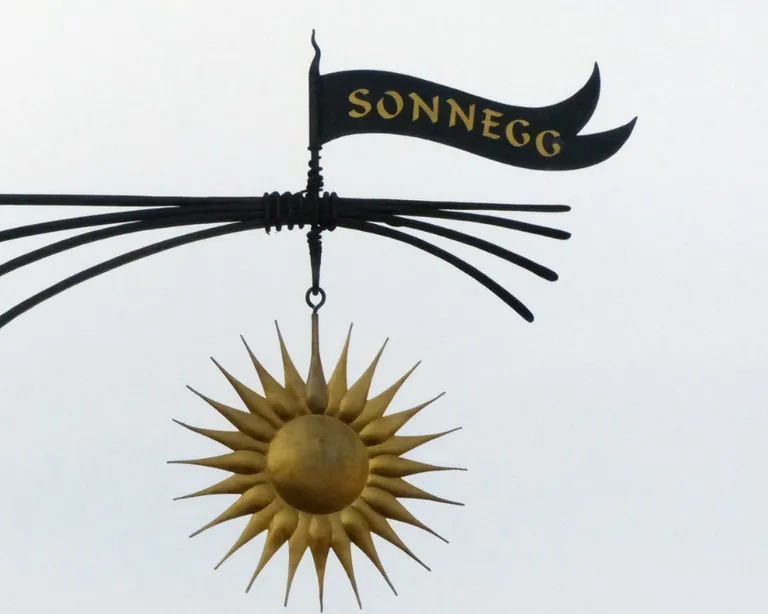 Schmiedeeisernes Schild mit einer goldenen Sonne und einer Fahne mit der Aufschrift «Sonneg» vor hellem Himmel.