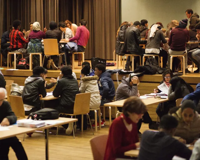 Viele Menschen sitzen in Gruppen an Tischen in einem grossen Saal und arbeiten gemeinsam oder unterhalten sich.