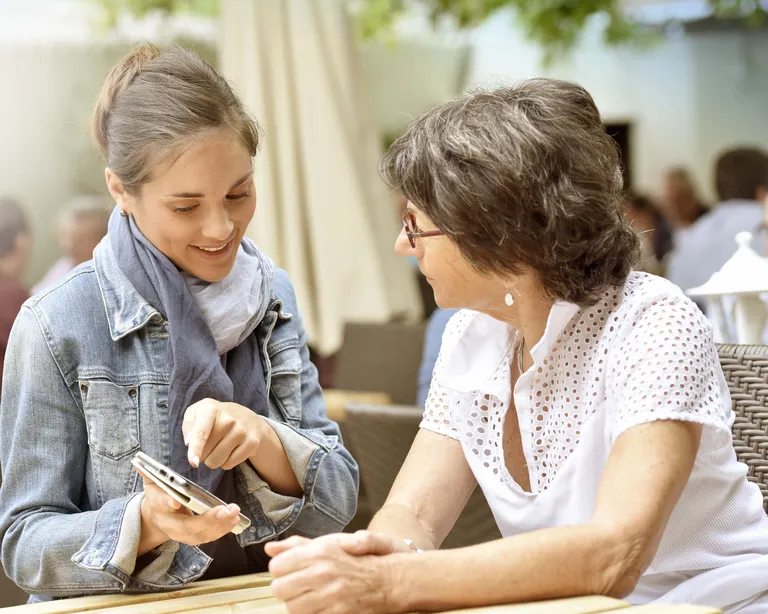 Eine junge Frau zeigt einer älteren Frau draussen an einem Tisch etwas auf ihrem Smartphone.