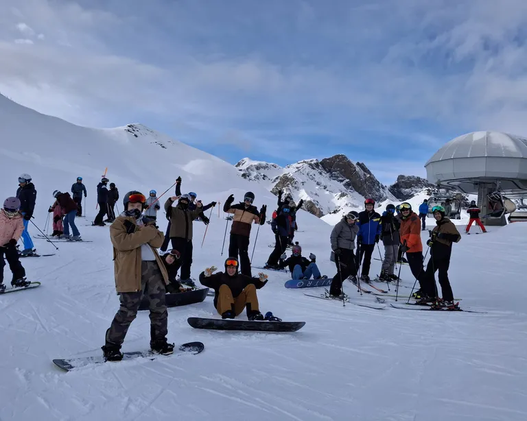 Jugendliche mit Snowboards und Skiern stehen am Snow Weekend auf einer verschneiten Piste bei sonnigem Wetter in den Bergen.