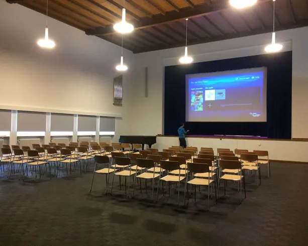 Ein leerer Seminarraum mit Stuhlreihen, grosser Leinwand und einer Person auf der Bühne vor einer Präsentation.