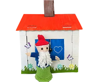 Ein kleiner Wichtel mit roter Mütze steht vor einem bunten, selbstgebastelten Häuschen mit rotem Dach und Blumen.