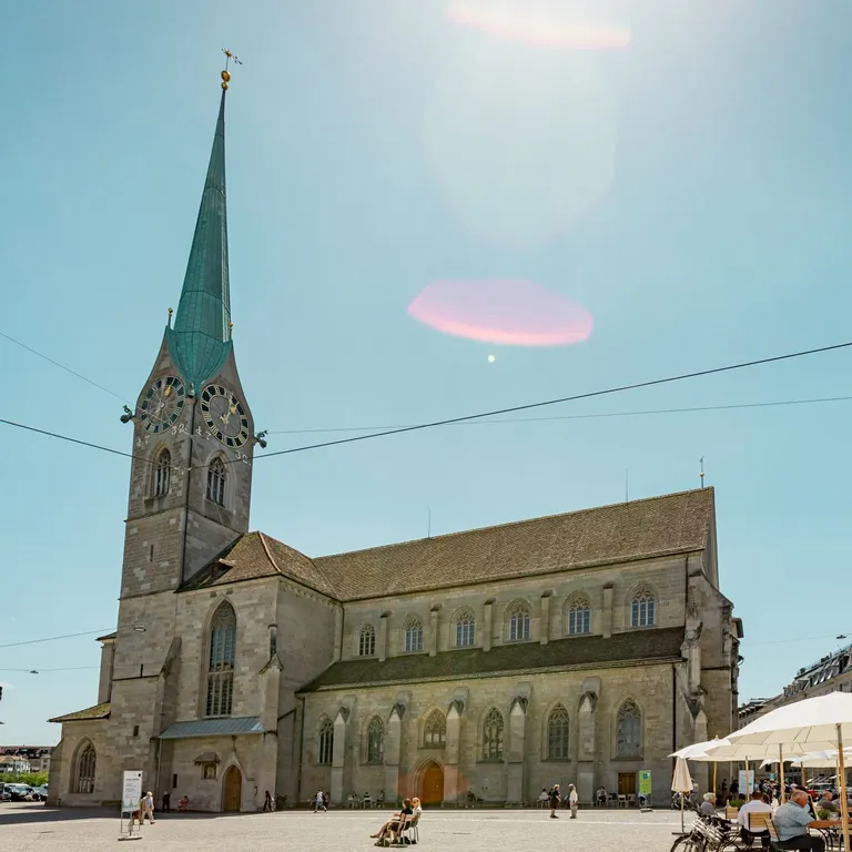 Fraumünster Kirche mit hohem Turm auf einem sonnigen Platz, Menschen sitzen unter Sonnenschirmen im Vordergrund.