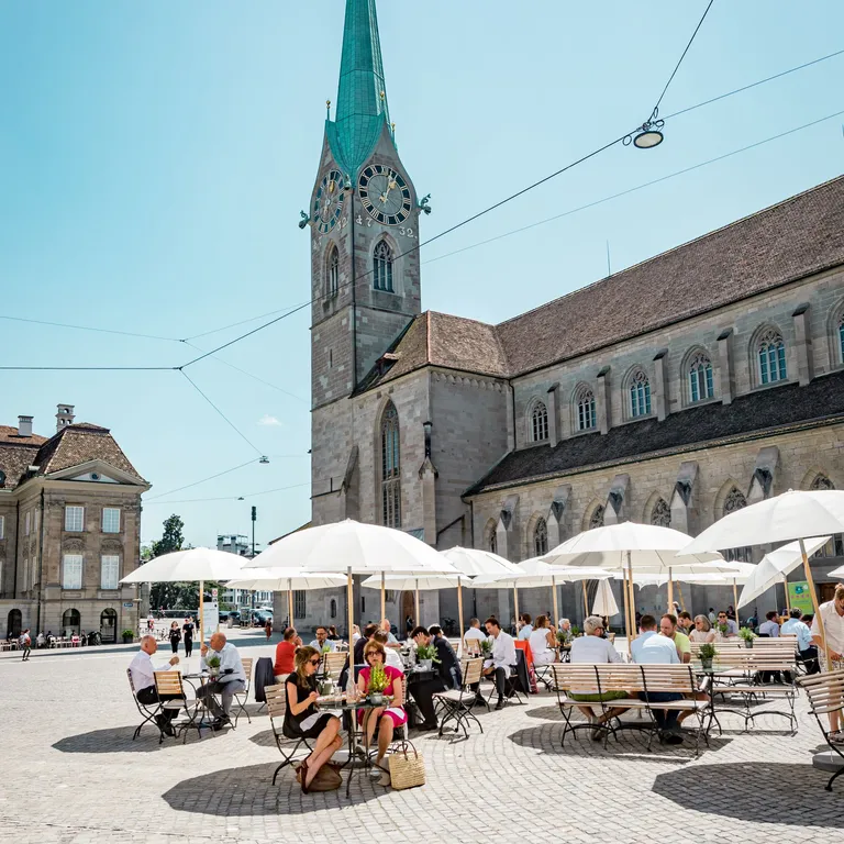 Menschen sitzen unter weißen Sonnenschirmen auf einem Platz vor der Kirche Fraumünster mit hohem Turm und grünem Dach.