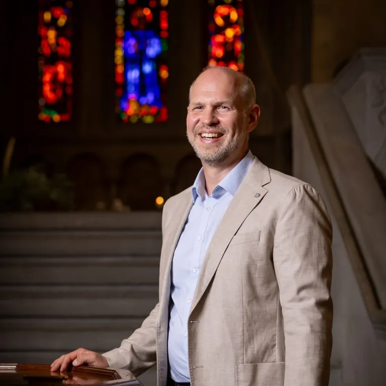 Pfarrer Christian Walti steht im hellen Sakko in einer Kirche vor einer Treppe, im Hintergrund sind bunte Kirchenfenster zu sehen.