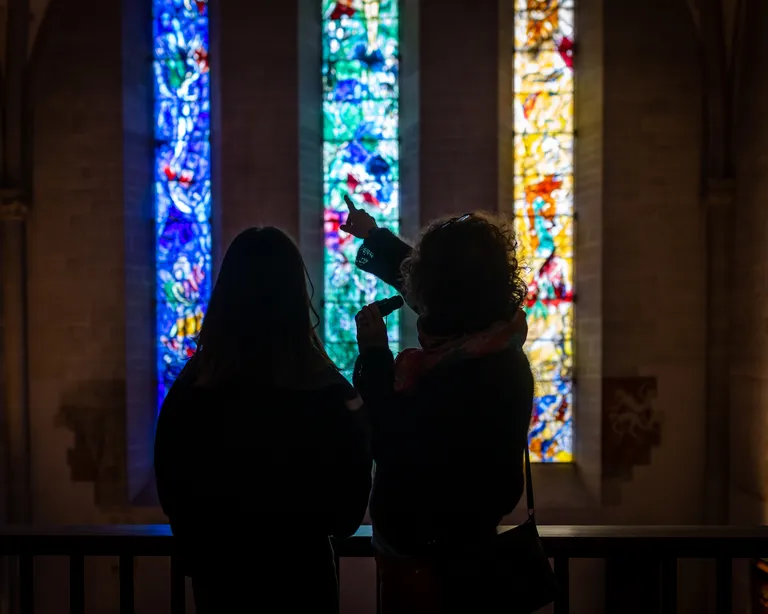 Zwei Personen stehen vor bunten Kirchenfenstern; eine Person zeigt mit der Hand auf das Glas.