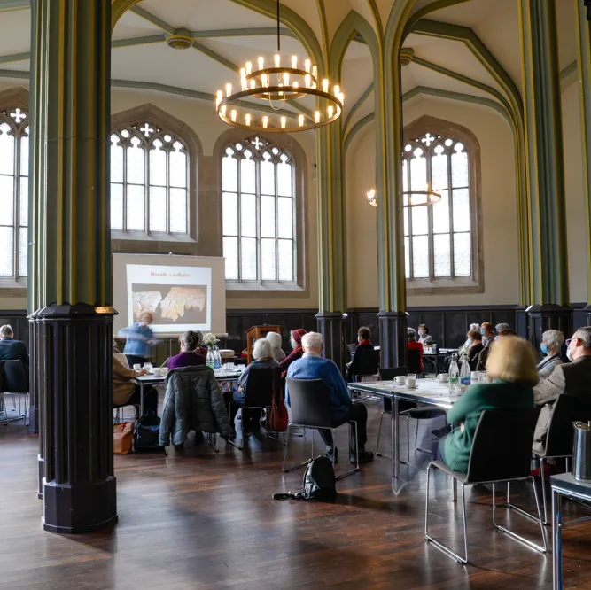Menschen sitzen an runden Tischen in einem großen, hellen Saal mit hohen Fenstern und Kronleuchtern.