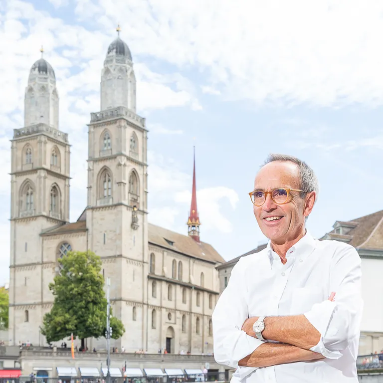 Ein lächelnder Mann steht vor dem Großmünster in Zürich bei Tageslicht, im Hintergrund sind historische Gebäude zu sehen.