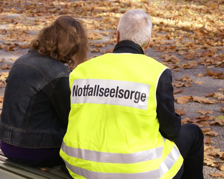 Zwei Personen sitzen auf einer Bank im Park, eine trägt eine gelbe Weste mit der Aufschrift «Notfallseelsorge».