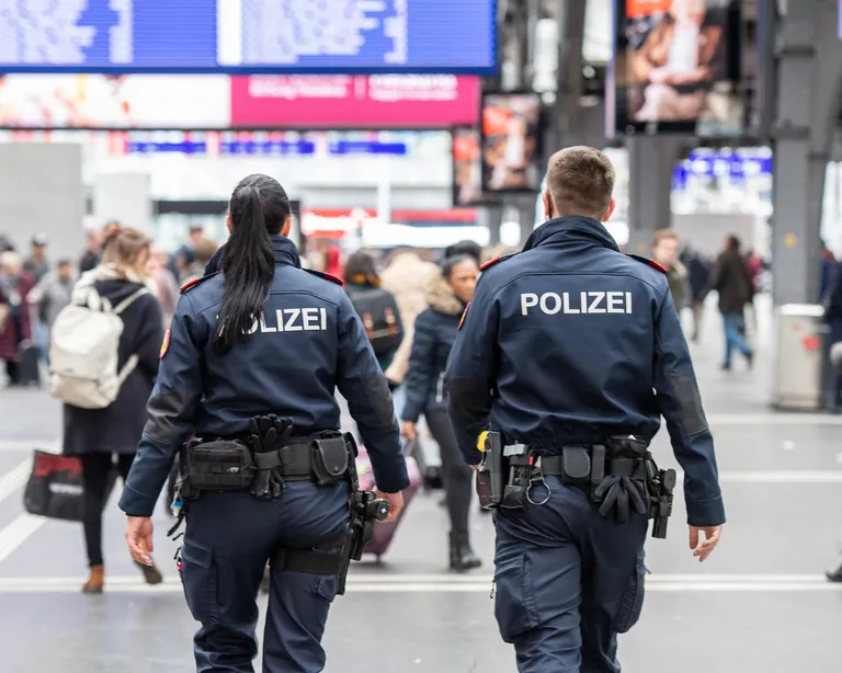 Zwei Polizisten gehen durch eine belebte Bahnhofshalle, umgeben von Passanten und Gepäck.
