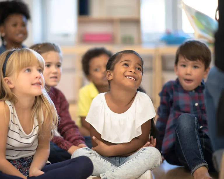 Mehrere Kinder sitzen lachend und aufmerksam im Kreis in einem Klassenraum oder Kindergarten.