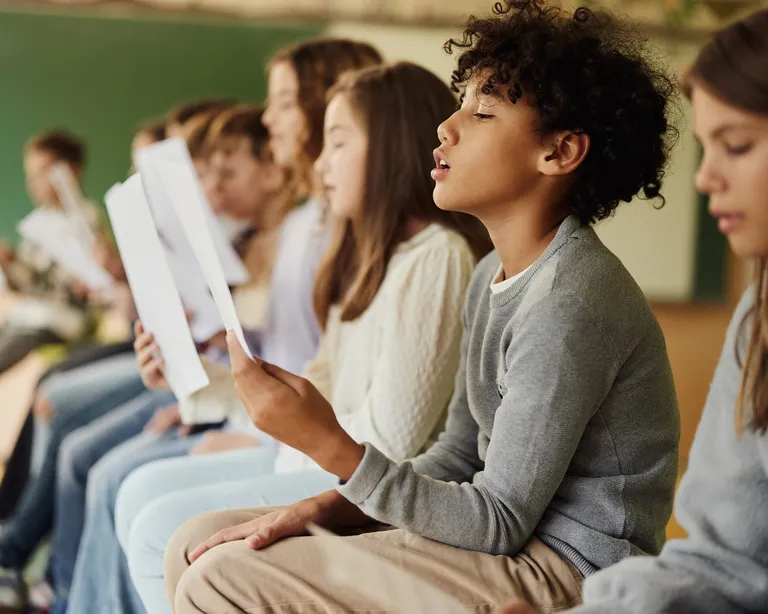Mehrere Kinder sitzen in einer Reihe und lesen gemeinsam aus Heften, vermutlich in einem Klassenzimmer.