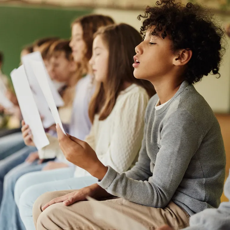 Mehrere Kinder sitzen in einer Reihe und lesen gemeinsam aus Heften, vermutlich in einem Klassenzimmer.