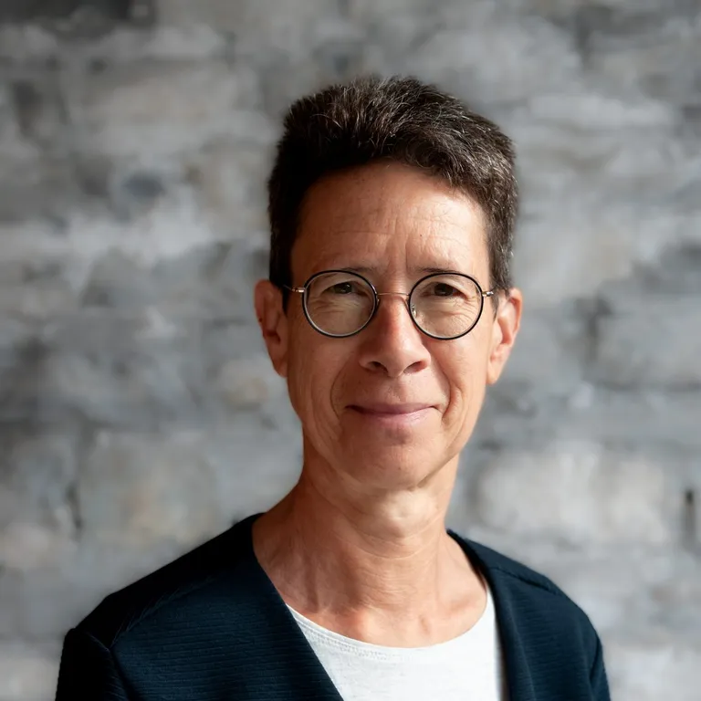 Porträt einer Person mit kurzem dunklem Haar, Brille und dunkler Jacke vor einem grauen, gemusterten Hintergrund.