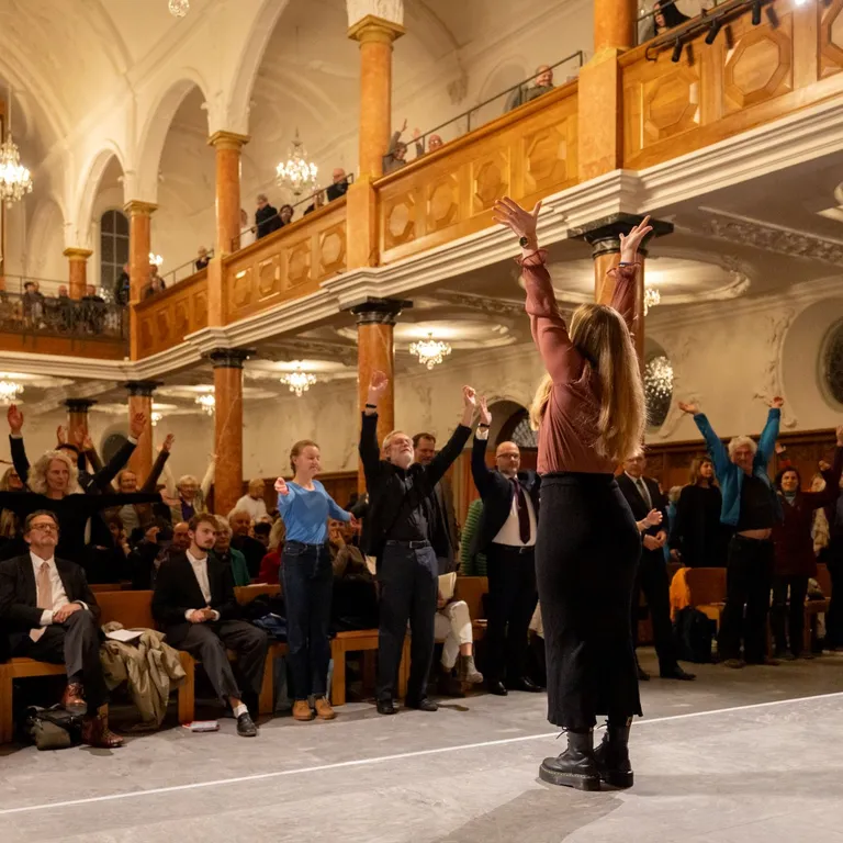 Eine Frau steht mit erhobenen Armen vor einer sitzenden und stehenden Menschenmenge in einer Kirche.