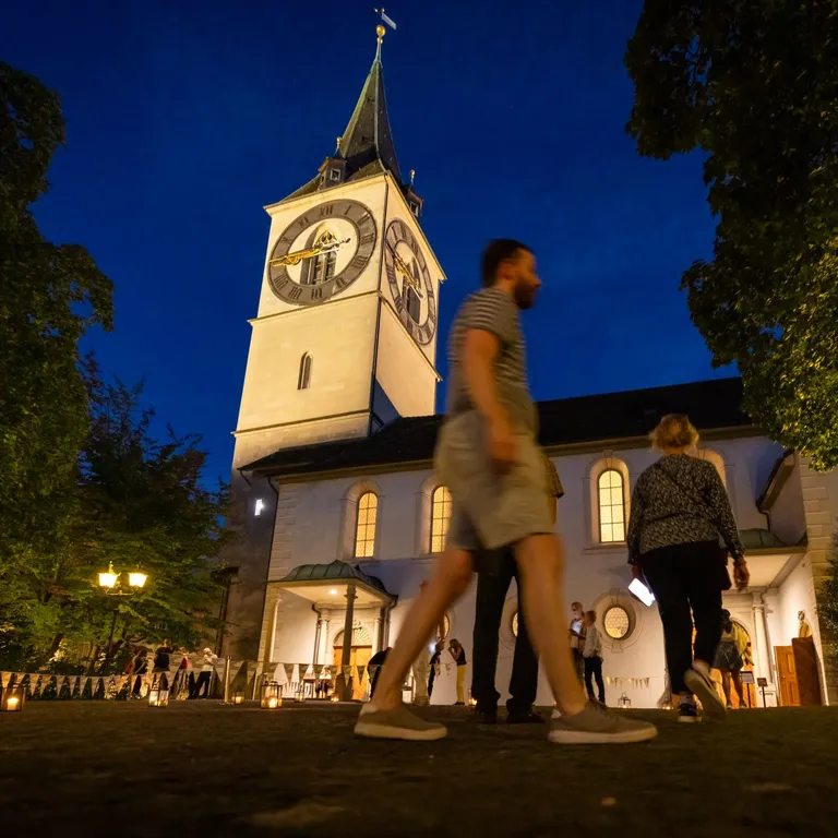 Mehrere Menschen gehen abends an einer beleuchteten Kirche mit grossem Uhrturm vorbei.