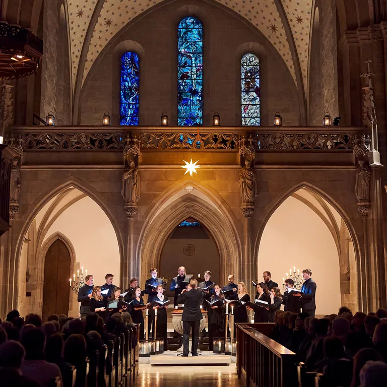 Ein Chor singt in einer beleuchteten Kirche mit hohen Gewölben und bunten Glasfenstern, Publikum sitzt vorne.