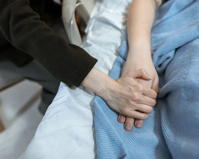 Zwei Menschen halten einander tröstend die Hand am Krankenbett, eine Person liegt unter einer blauen Decke.
