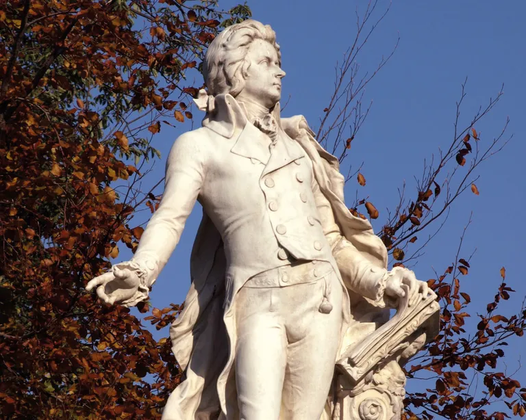 Weiße Statue eines Mannes im eleganten Anzug, im Freien, mit Bäumen und blauem Himmel im Hintergrund.