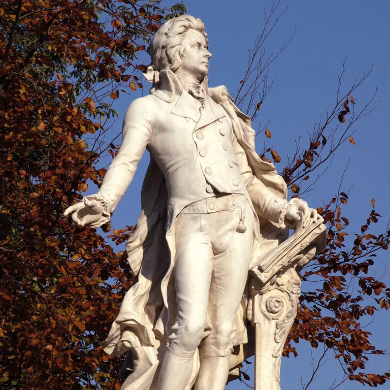 Weiße Statue eines Mannes im eleganten Anzug, im Freien, mit Bäumen und blauem Himmel im Hintergrund.