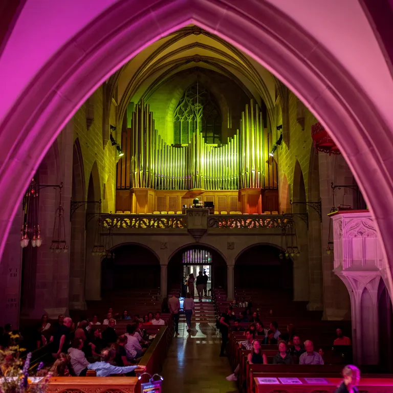 Blick auf die Fraumünsterorgel auf der Empore bei Nacht und farbig beleuchteter Kirche in Pink und Gelb.